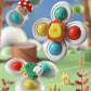 A colorful children's toy fidget spinner shown in a playful outdoor-style scene with soft-focus background elements. The main spinner has a white body, a yellow lion face in the center with red petals, and four round push buttons in teal, yellow, and red.