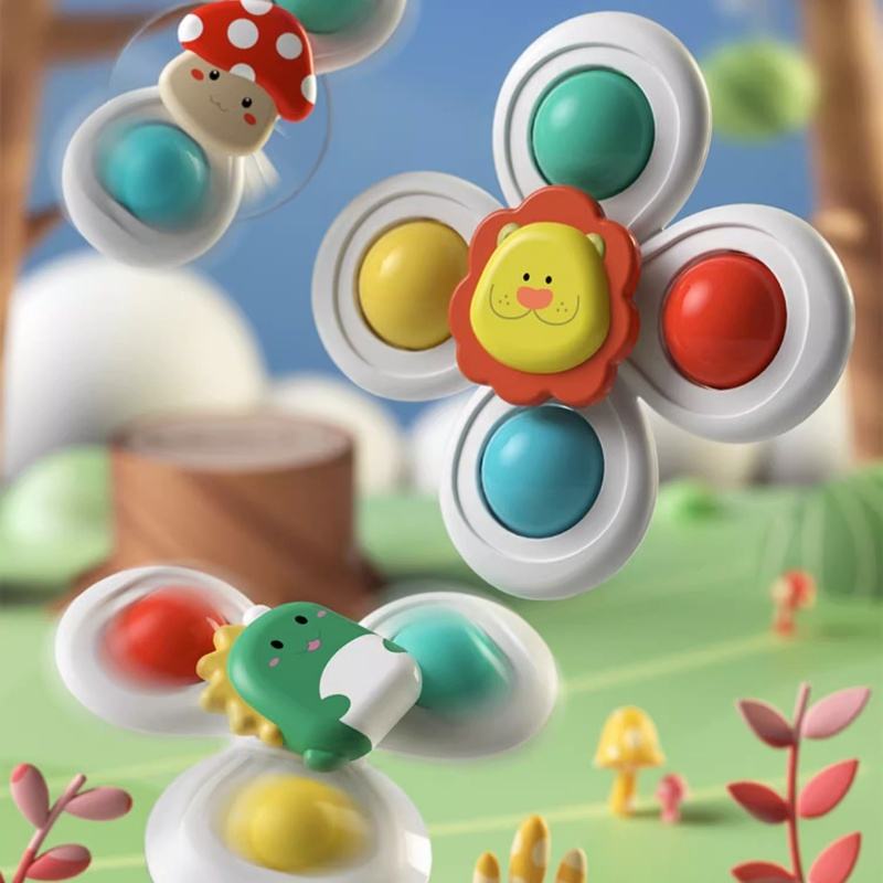 A colorful children's toy fidget spinner shown in a playful outdoor-style scene with soft-focus background elements. The main spinner has a white body, a yellow lion face in the center with red petals, and four round push buttons in teal, yellow, and red.