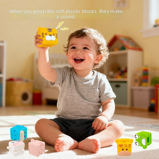 A smiling toddler sits on a carpet in a bright playroom, holding up a yellow soft plastic block while other colorful blocks are scattered around them. Text at the top reads, 'When you pinch the soft plastic blocks, they make a sound.